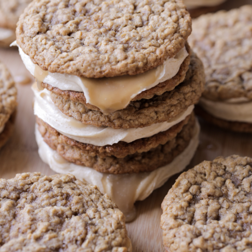 Three sourdough discard browned butter caramel apple oatmeal cream pies sit on top of each other. They have a brown sugar cream cheese frosting filling and are surrounded by several oatmeal cream pies.