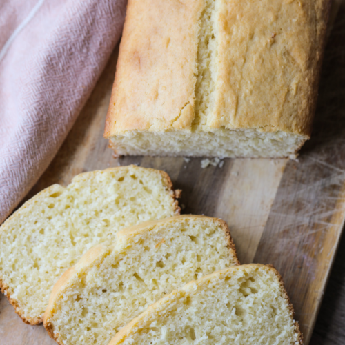 Three slices of sourdough discard vanilla pound cake is shown next to the rest of the loaf on a wooden cutting board.