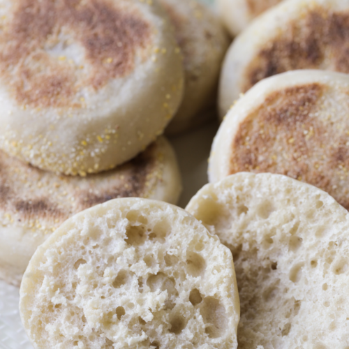 Several sourdough english muffins are shown golden brown and perfectly round. One english muffin is shown cut in half where you can see the nooks and crannies.