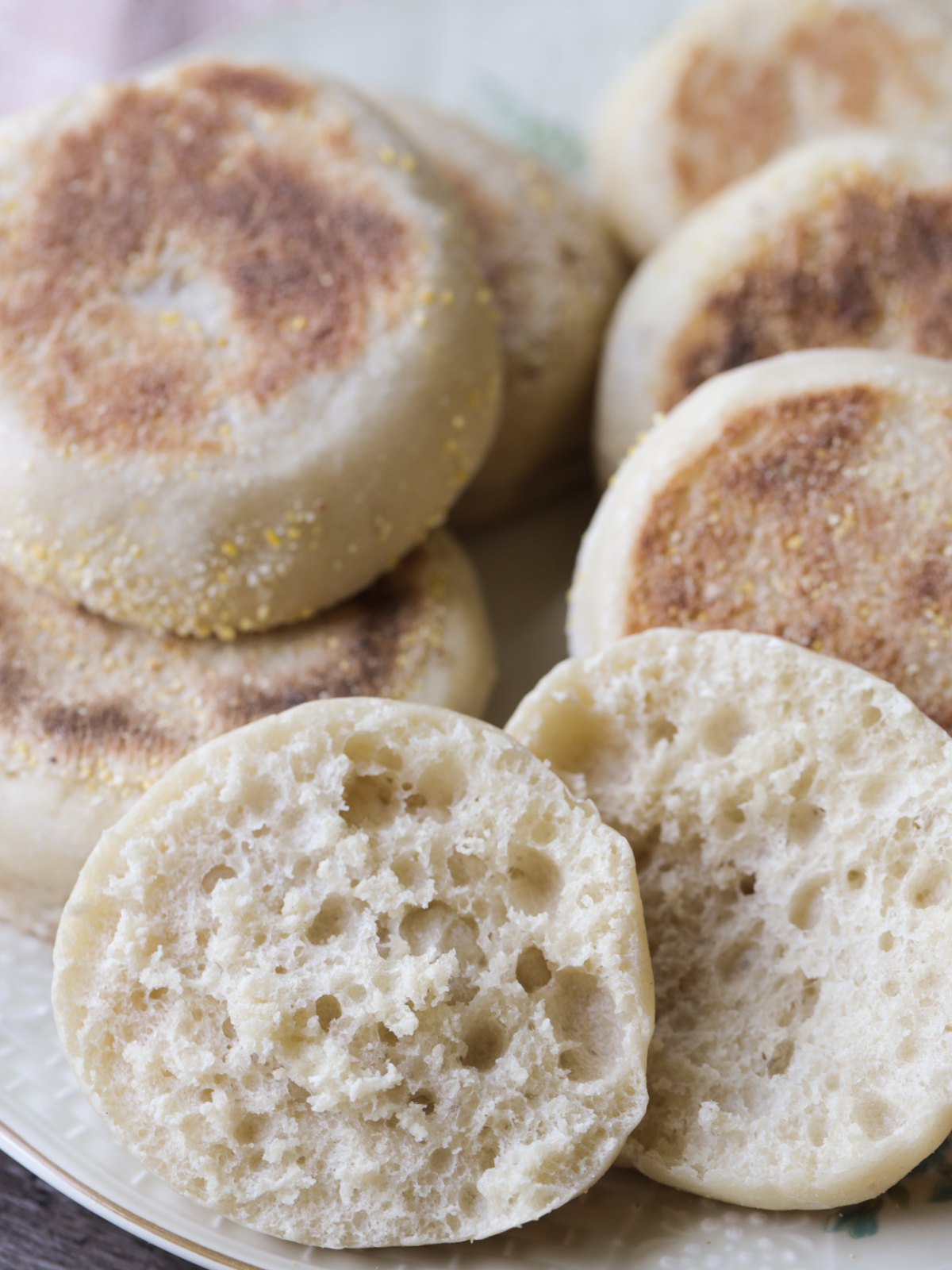 Several sourdough english muffins are shown golden brown and perfectly round. One english muffin is shown cut in half where you can see the nooks and crannies.