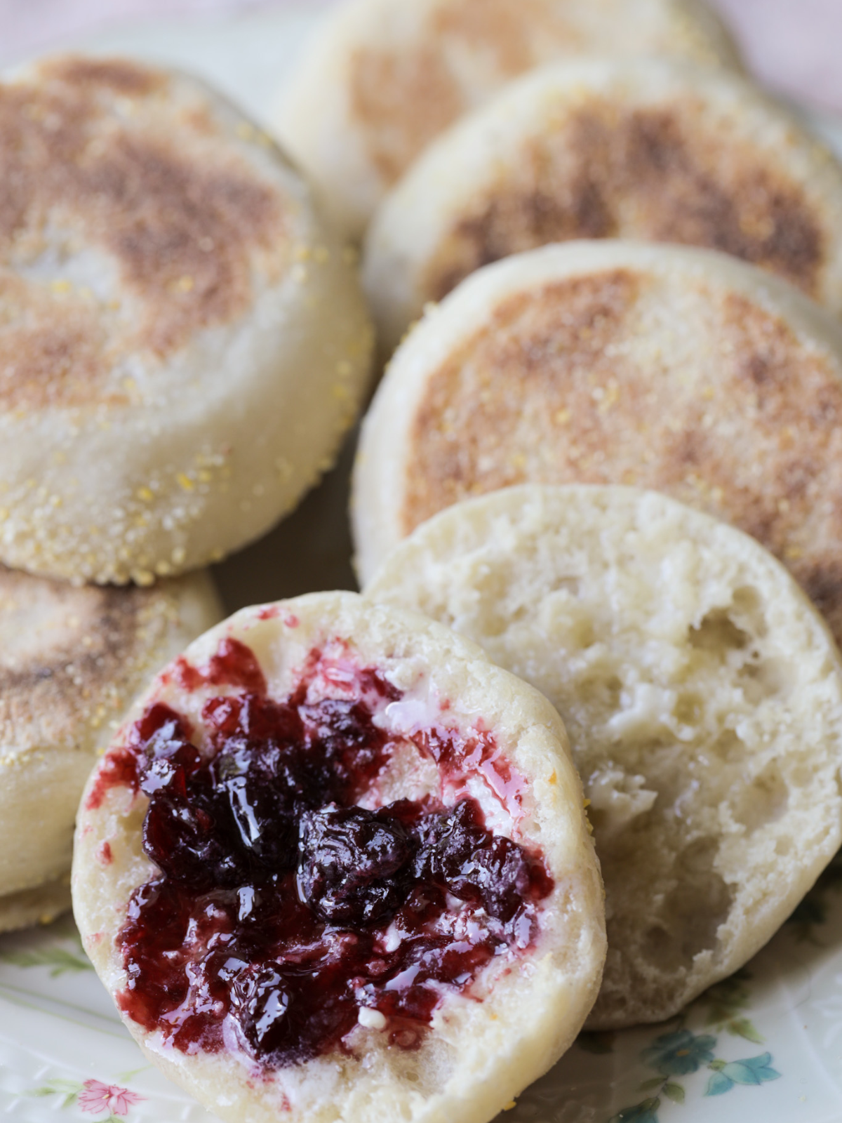 A 100% sourdough English muffin is shown with jam on it. The picture shows several other freshly made English muffins in the background.