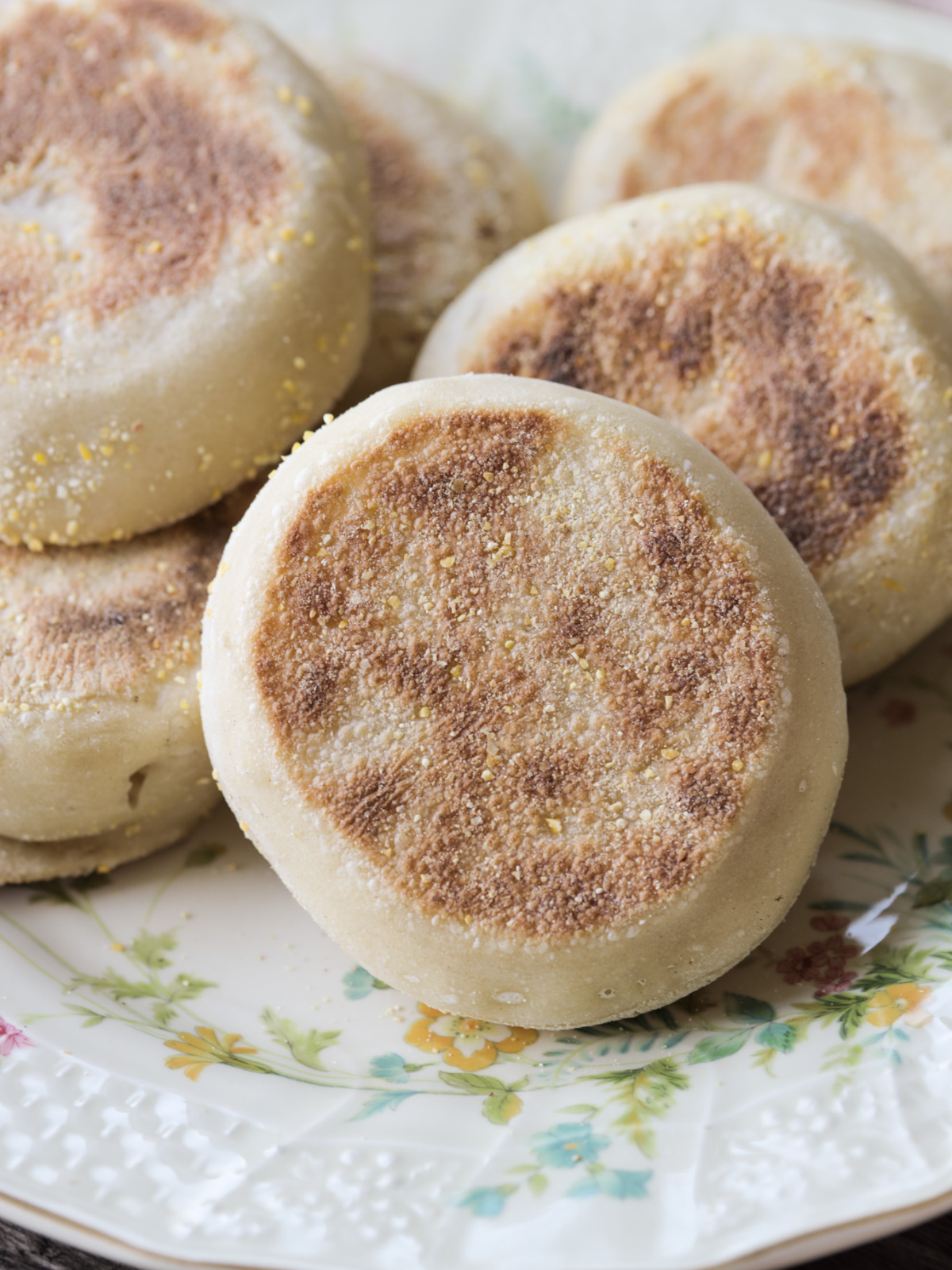 A closeup of freshly made sourdough English muffins sit on a floral plate. Golden brown on both sides and a sprinkling of cornmeal over them all.