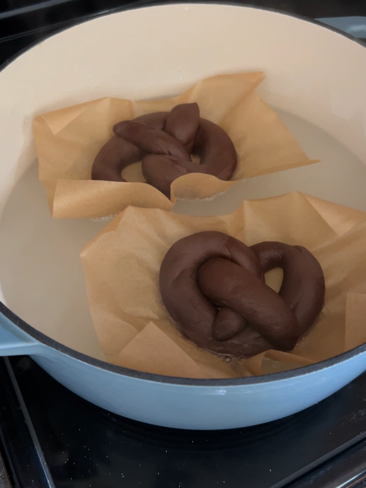 Two sourdough chocolate pretzels are shown boiling in the baking soda bath.