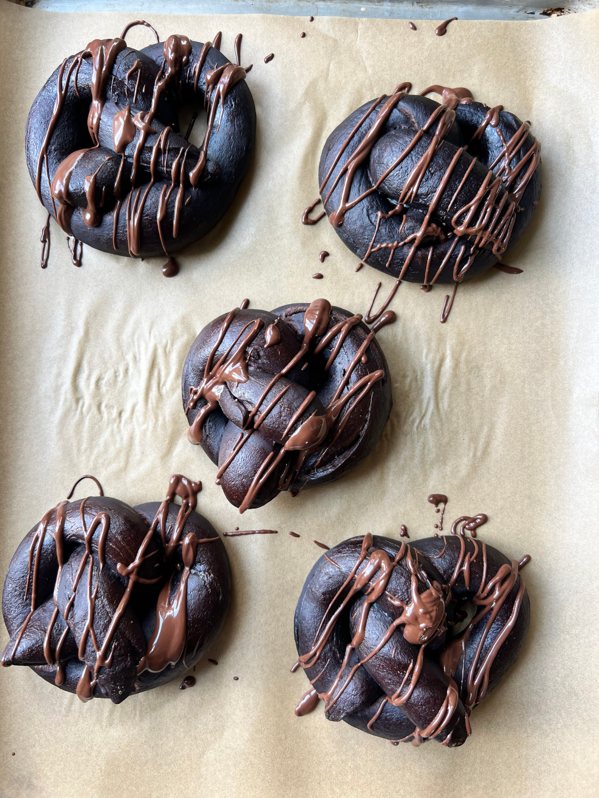 Five sourdough chocolate soft pretzels rest on a parchment paper-lined baking sheet. They are drizzled with chocolate.