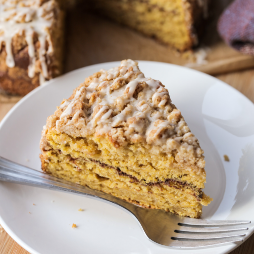 A slice of sourdough discard brown butter pumpkin swirl coffee cake is shown on a white plate with a fork. The rest of the coffee cake sits in the background.