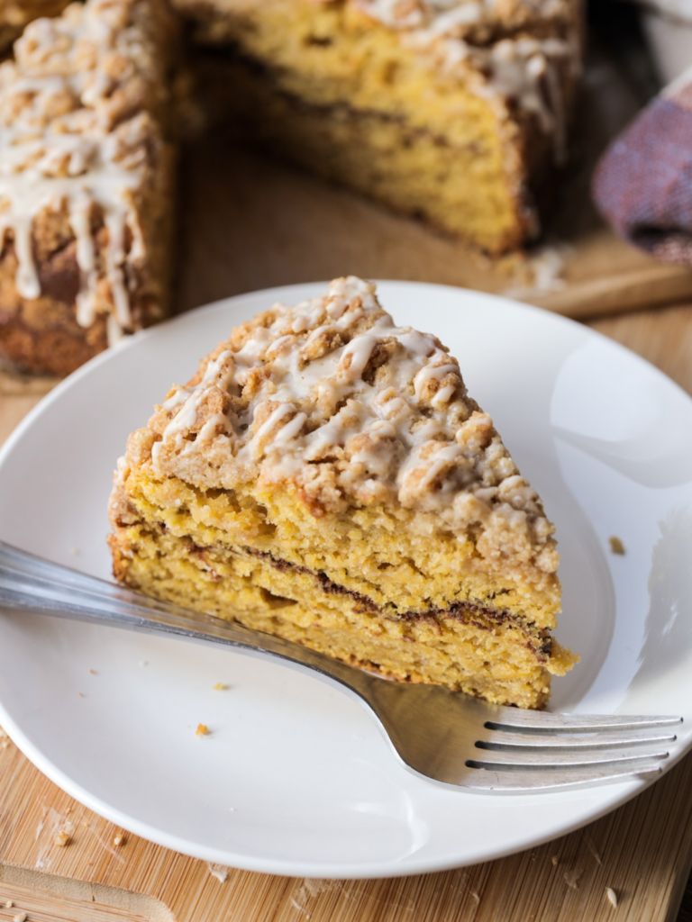 A slice of sourdough discard brown butter pumpkin swirl coffee cake is shown on a white plate with a fork. The rest of the coffee cake sits in the background.