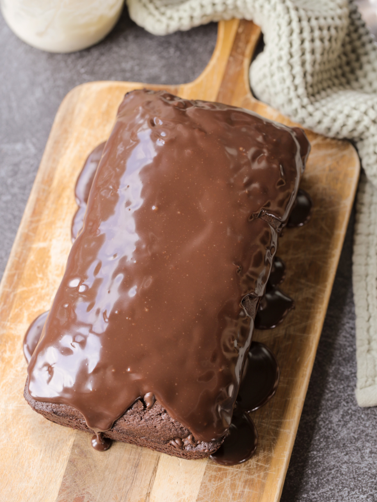 The sourdough brownie loaf rests on a wooden cutting board. Chocolate ganache has been poured over it.