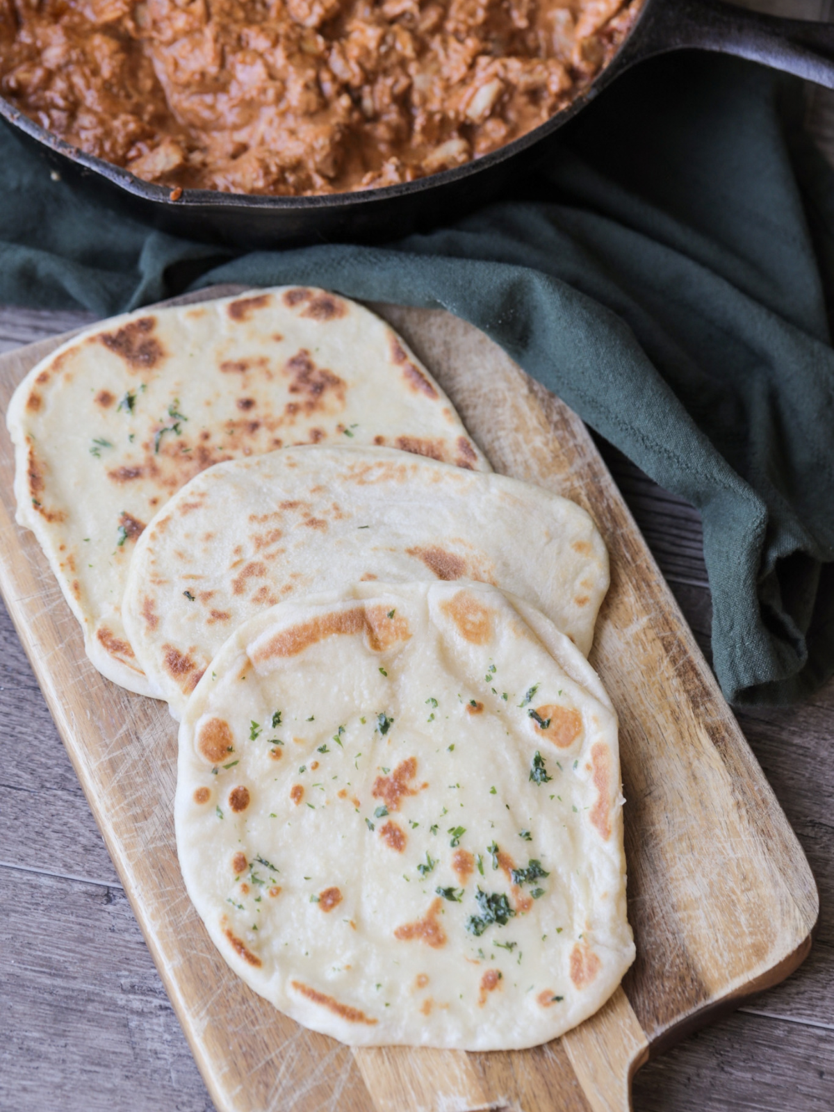 Homemade sourdough discard naan sits on a wooden cutting board. Butter chicken sits in a cast iron skillet behind the naan.