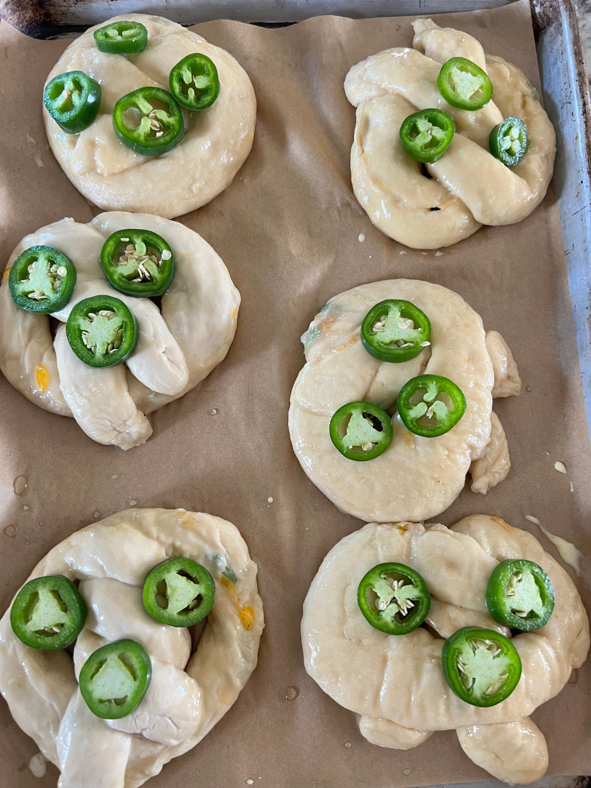 The freshly boiled and egg wash brushed sourdough jalapeno and cheddar pretzels are shown on a parchment paper lined baking sheet before they bake.
