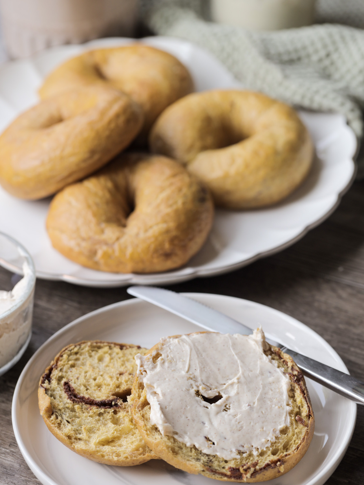 A sourdough pumpkin cinnamon swirl bagel is shown cut in half on a plate with a cinnamon maple cream cheese spread over it. Several sourdough pumpkin cinnamon swirl bagels are shown on a platter in the background.