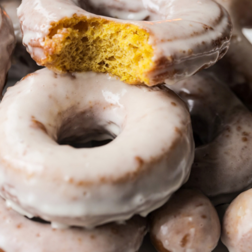 A closeup of two sourdough pumpkin donuts with maple glaze are shown. One donuts has a bite taken out so you can see the fluffy interior.
