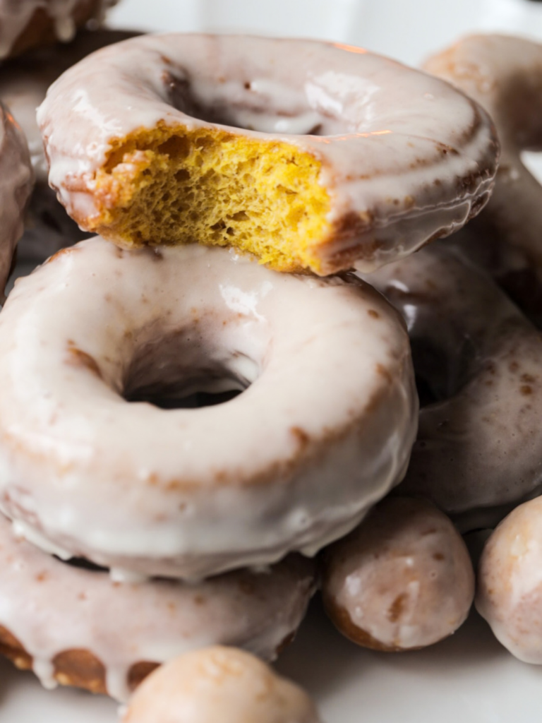 A closeup of two sourdough pumpkin donuts with maple glaze are shown. One donuts has a bite taken out so you can see the fluffy interior.