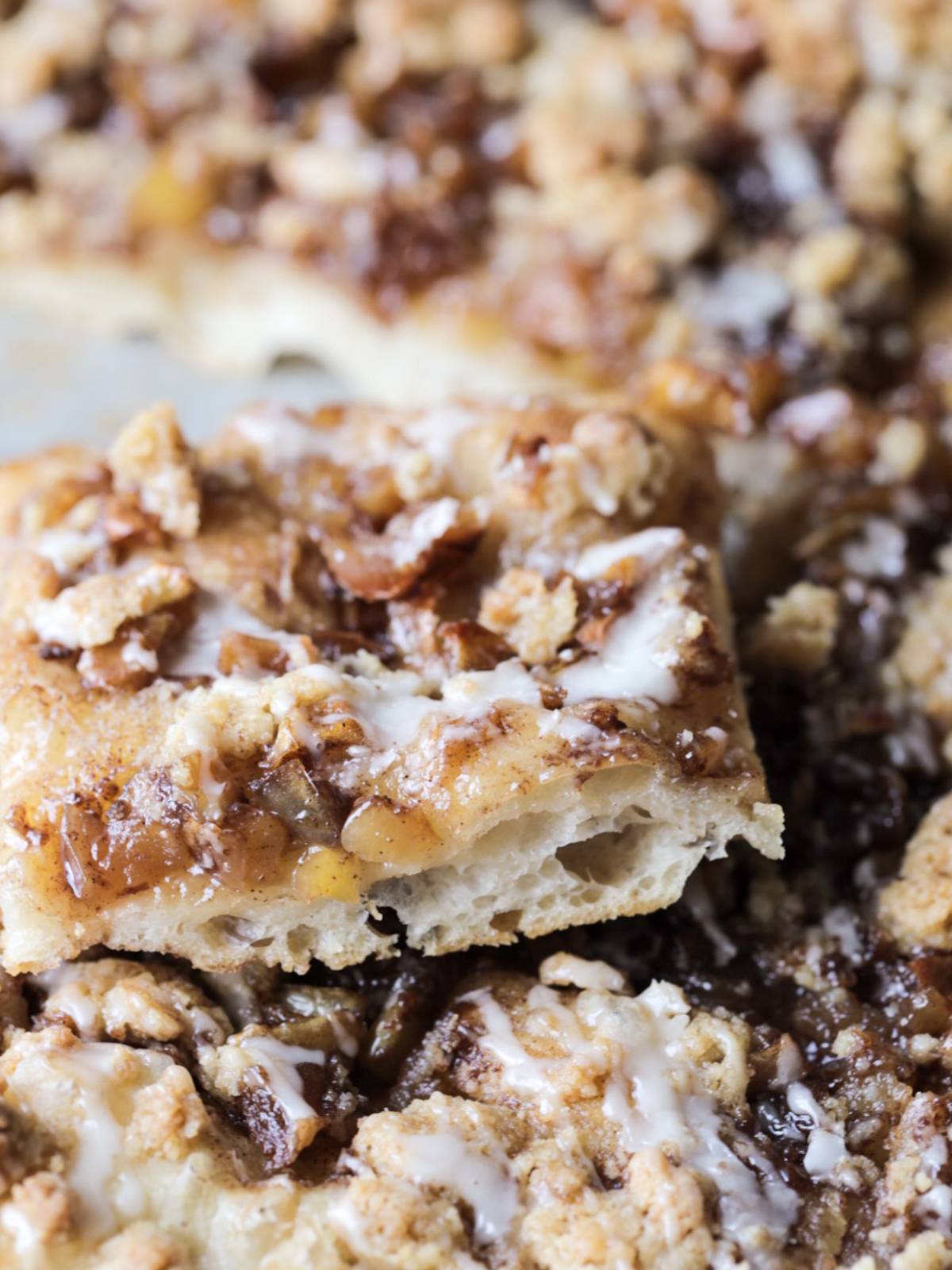 A slice of sourdough apple crisp focaccia is shown with an airy crumb. Vanilla icing is drizzled and a simple crumble is on top.