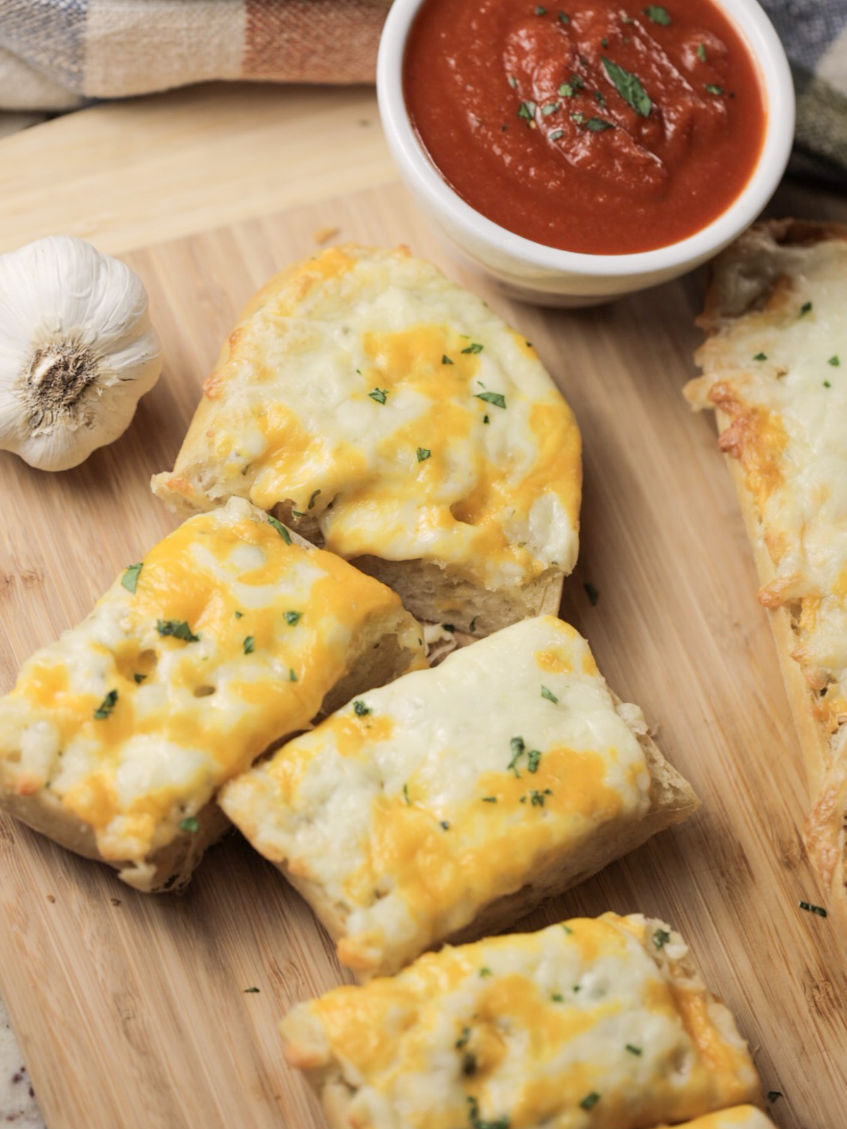 Several slices of cheesy sourdough garlic french bread is shown on a wooden cutting board with a bowl of marinara sauce in the background.