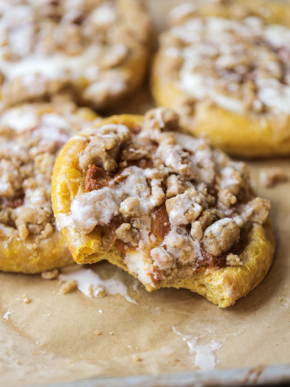 A Sourdough brioche pumpkin kolache with brown sugar crumble and vanilla icing is shown on a parchment paper lined baking sheet with two bites taken out of it.